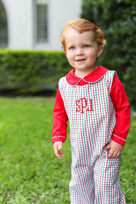 A toddler standing outdoors on grass wearing a red and white gingham overall jumpsuit with a red long-sleeve shirt underneath and red monogram lettering on the chest.