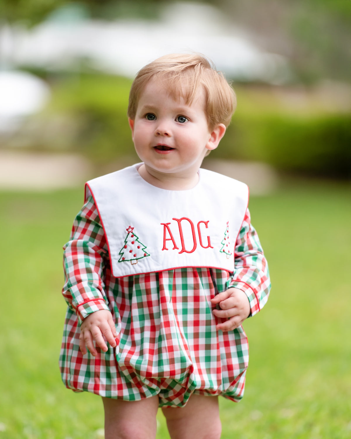 A toddler standing outdoors on grass wearing a red, green, and white plaid romper with a white bib featuring red monogram letters and small Christmas tree motifs.