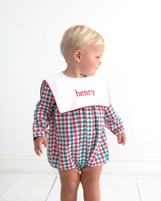 A toddler stands in profile wearing a red, green, and white gingham long-sleeve romper with a white bib featuring the name 'henry' in red script.