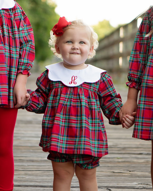 A toddler stands on a wooden boardwalk wearing a red plaid dress with a white collar and a red monogrammed 'H', holding hands with two other children in matching outfits. One child wears a red bow in her hair.