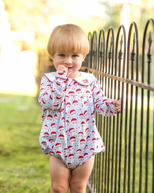 A toddler stands outdoors in front of a black metal fence, wearing a white long-sleeve romper covered in a red Santa Claus hat pattern with a white collar.