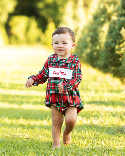 A toddler walking barefoot on a grassy lawn outdoors, wearing a red and green plaid romper dress with a white name tag that reads 'hughes'.