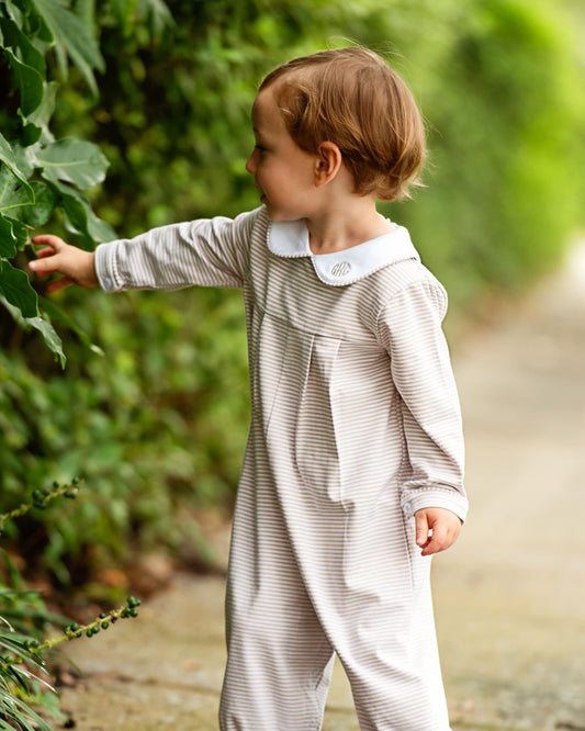 A toddler wearing a long-sleeve striped romper with a white collar stands outdoors on a path, reaching toward green leaves.