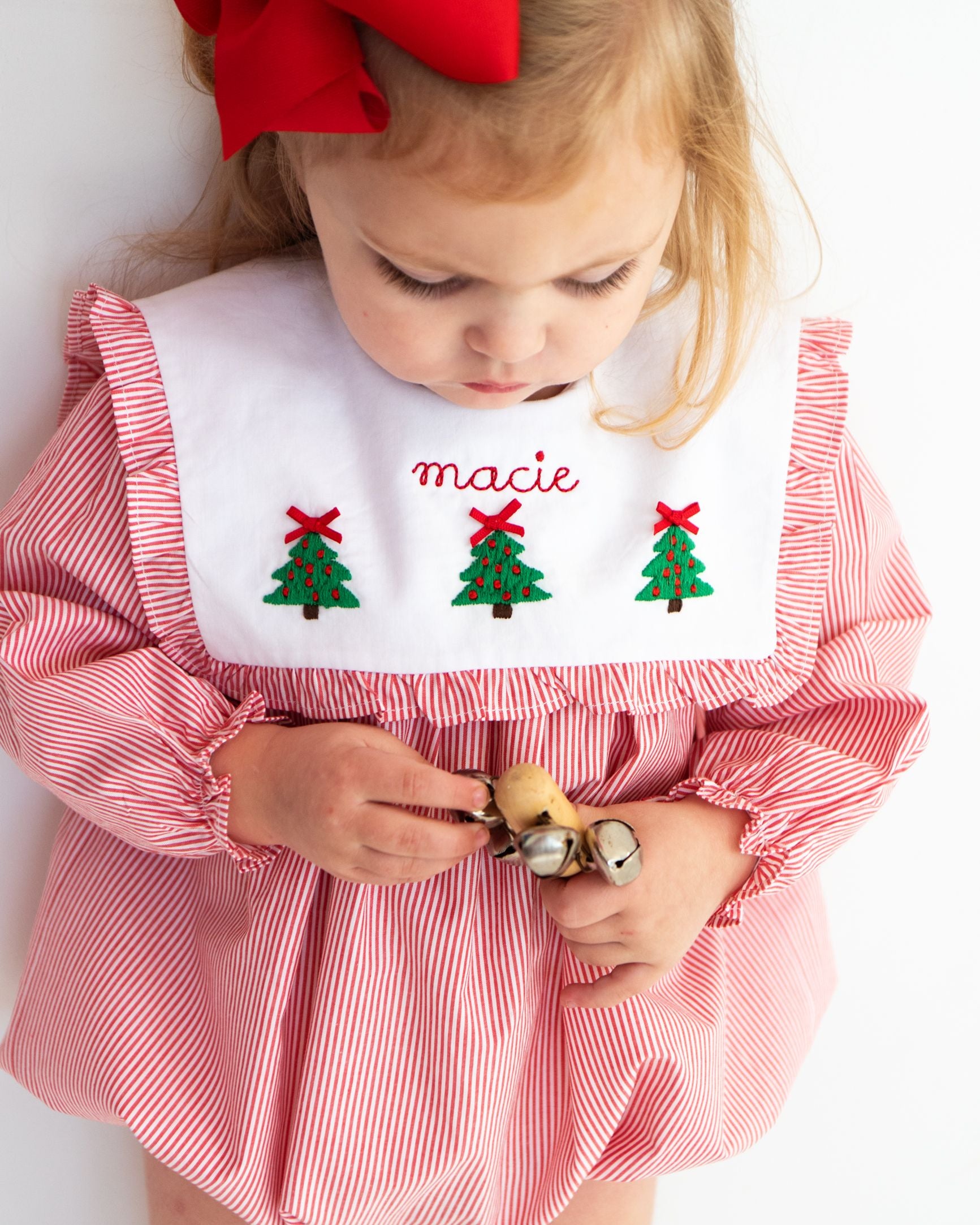 A toddler wearing a red and white striped long-sleeve dress with a white bib personalized with the name 'macie' and embroidered Christmas trees. The child is holding small bells against a clean white background.