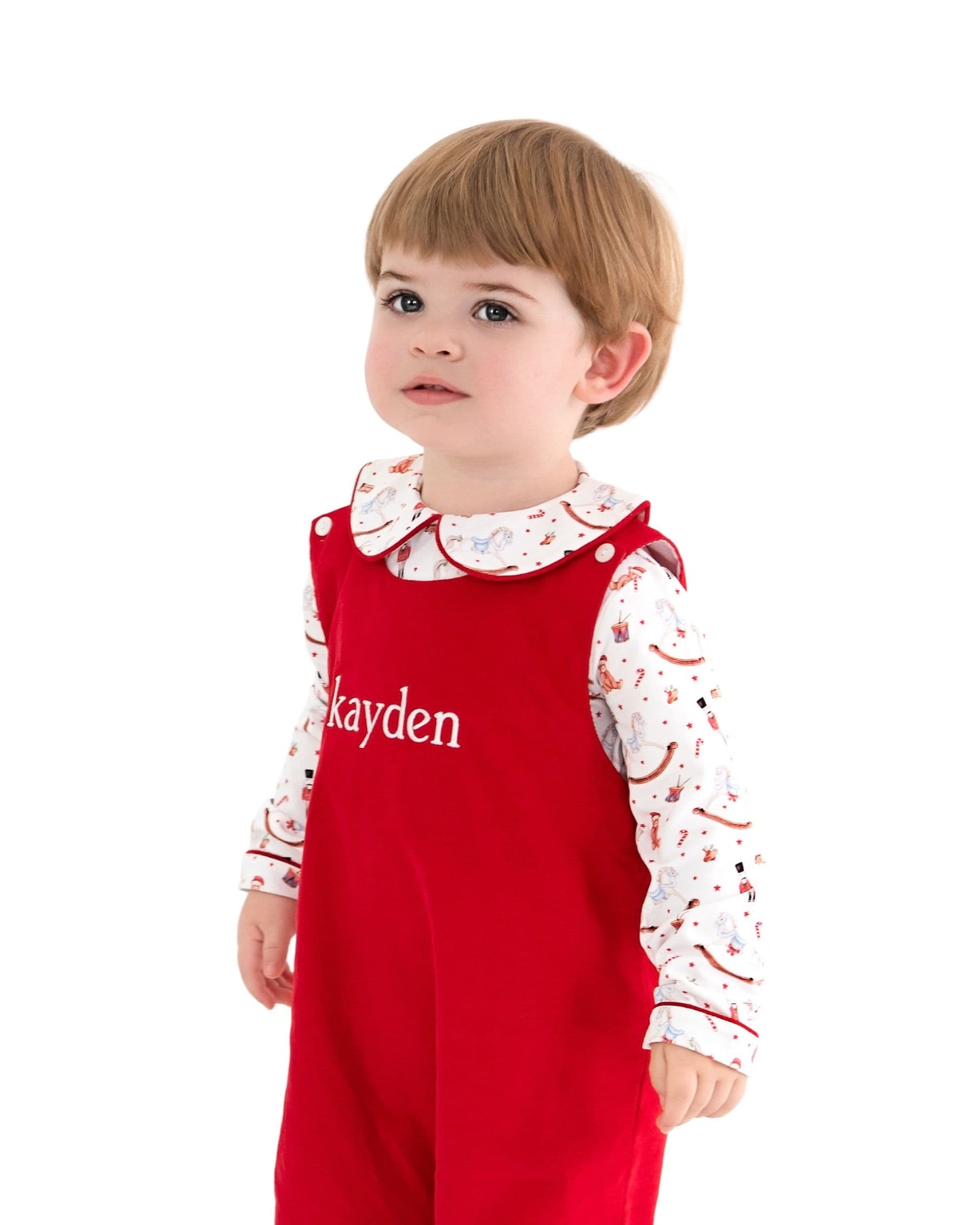 A toddler wearing a red overall-style romper over a white long-sleeve shirt with a multicolor holiday print and a white Peter Pan collar. The red romper has the name 'kayden' printed in white on the chest.
