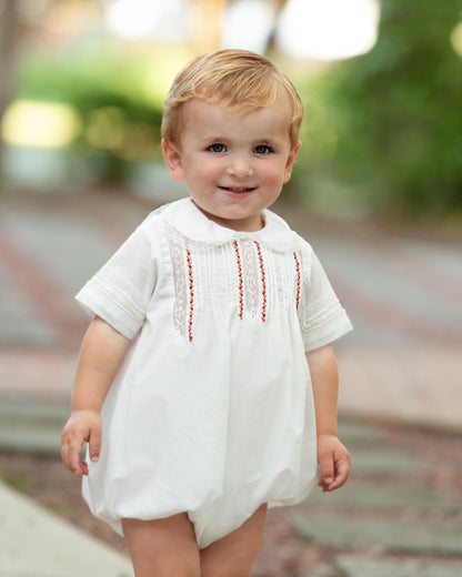 A toddler wearing a white short-sleeve romper with red embroidered vertical stripes on the chest, standing outdoors on a blurred path.
