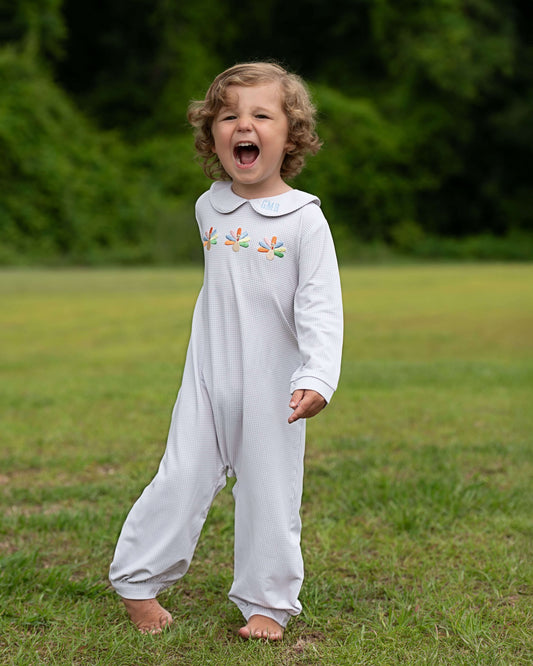 A toddler with curly light brown hair stands barefoot on a grassy lawn outdoors, wearing a white long-sleeve footless baby jumpsuit with colorful embroidered leaf motifs on the chest.