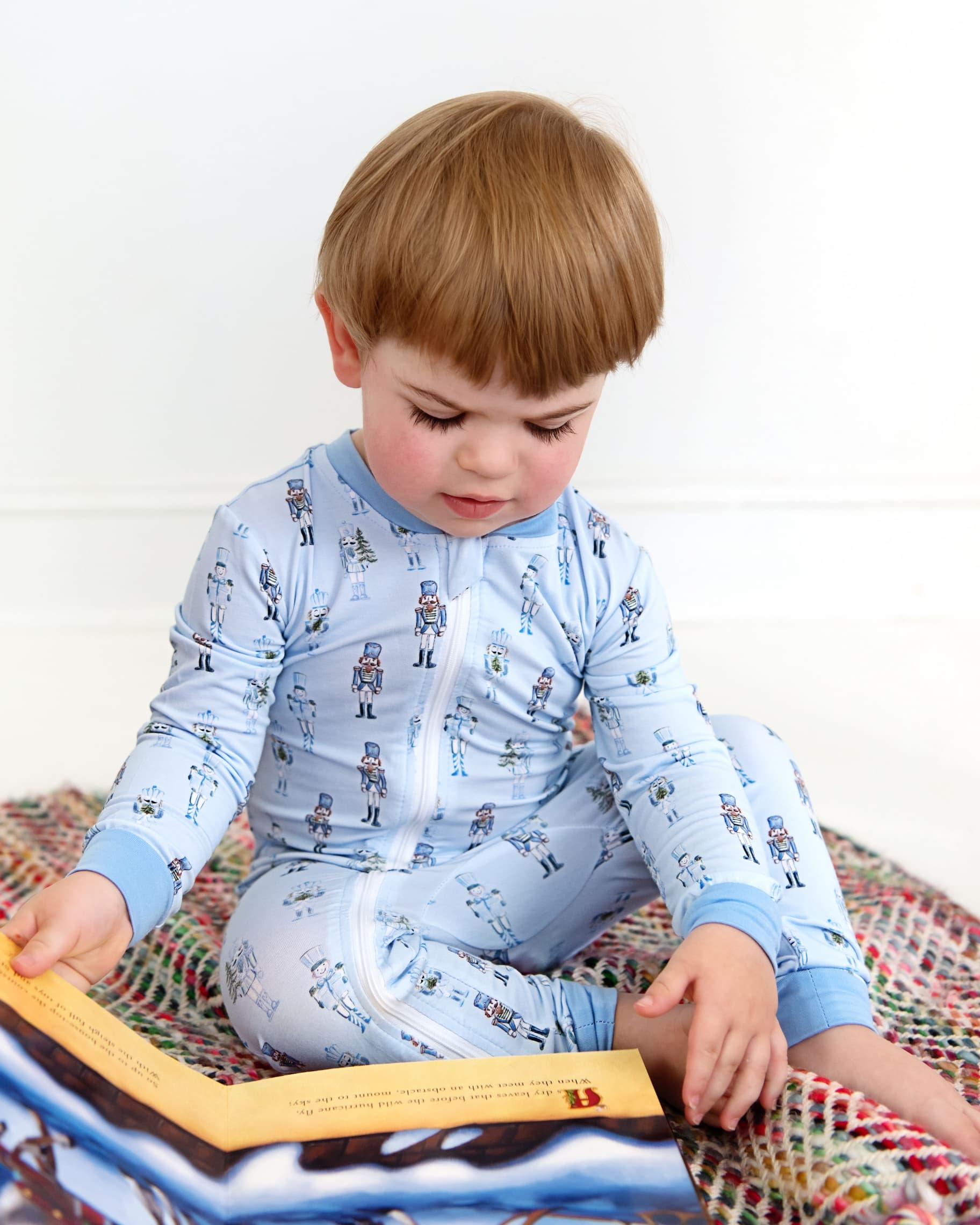 A toddler with light brown hair sits on a colorful woven blanket, reading a picture book. The child is wearing a light blue long-sleeve zip-up pajama set with a small repeating astronaut pattern.