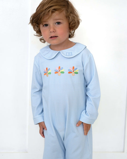 A toddler with wavy light brown hair wearing a light blue gingham long-sleeve romper with a Peter Pan collar and embroidered dragonflies on the chest, standing against a white paneled wall.