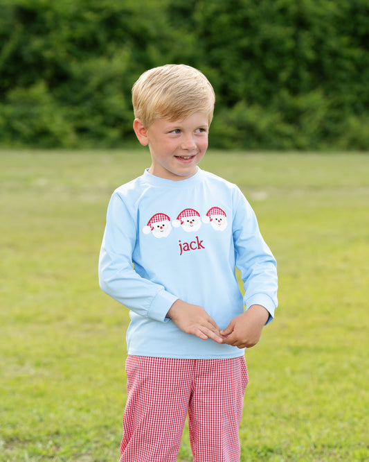 A young blond boy standing on a grassy lawn wearing a light blue long-sleeve shirt with three red-and-white striped cap graphics and the name 'jack' in red, paired with red-and-white gingham pants.