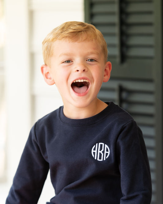 A young blond boy with blue eyes wearing a navy blue long-sleeve crewneck sweatshirt with a white circular monogram logo on the chest, captured in a joyful expression.
