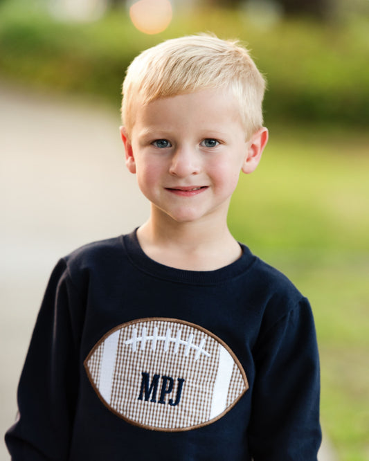 A young blonde boy smiling at the camera, wearing a navy blue long-sleeve sweatshirt with a football graphic on the front featuring the letters 'MPJ'.