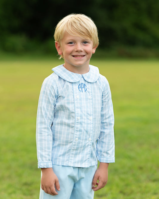 A young blonde boy smiling outdoors in a light blue and white plaid long-sleeve shirt with a Peter Pan collar and matching light blue trousers. The shirt has a blue monogram on the chest.