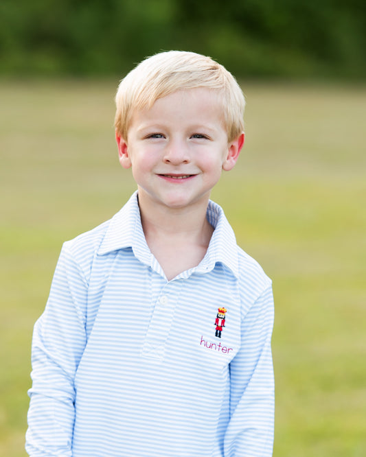A young blonde boy smiling outdoors in a light blue and white striped polo shirt with a small embroidered nutcracker and the name 'hunter' on the chest pocket.
