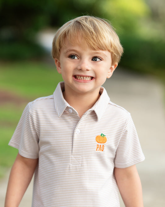 A young blonde boy smiling outdoors, wearing a light beige and white striped short-sleeve polo shirt with an orange and green 'PAC' logo on the left chest.