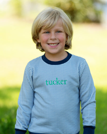 A young blonde boy smiling outdoors, wearing a light blue and white gingham long-sleeve shirt with dark navy ribbed collar, cuffs, and hem, and the name 'macker' printed in teal across the chest.