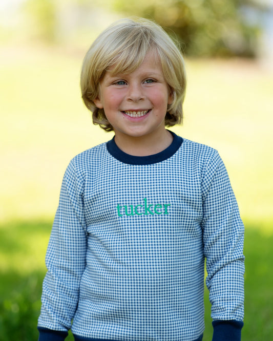 A young blonde boy smiling outdoors, wearing a light blue and white gingham long-sleeve shirt with dark navy ribbed collar, cuffs, and hem, and the name 'macker' printed in teal across the chest.