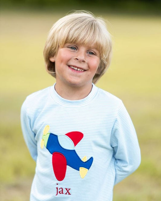 A young blonde boy smiling outdoors, wearing a light blue long-sleeve shirt with a colorful airplane graphic and the name 'jax' printed below it.
