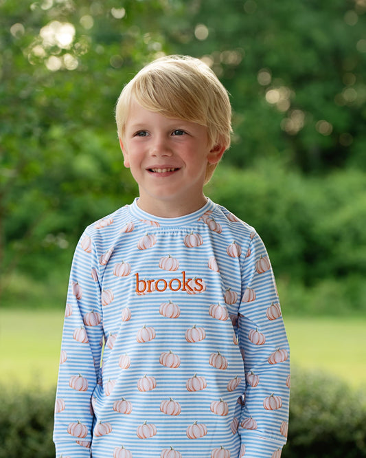 A young blonde boy smiling outdoors, wearing a light blue long-sleeve shirt with thin horizontal stripes, a pumpkin pattern, and the name 'brooks' printed in orange across the chest.