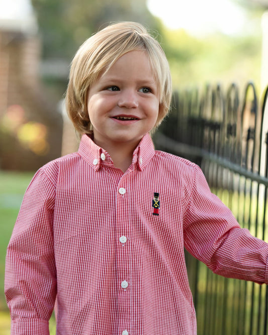 A young blonde boy smiling outdoors, wearing a red and white gingham long-sleeve button-down shirt with a small embroidered toy soldier logo on the chest.