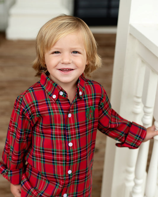 A young blonde boy smiling while wearing a red plaid long-sleeve button-down shirt with green and black stripes, standing next to a white wooden staircase.