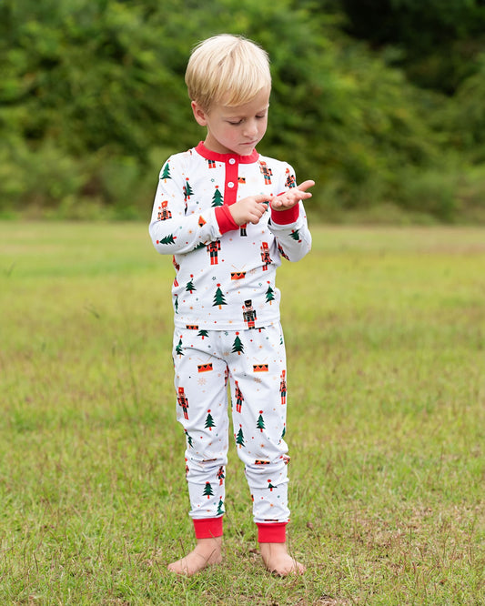A young blonde boy stands barefoot on a grassy field wearing a white long-sleeve pajama set with a red collar and cuffs, decorated with a nutcracker and Christmas tree pattern.