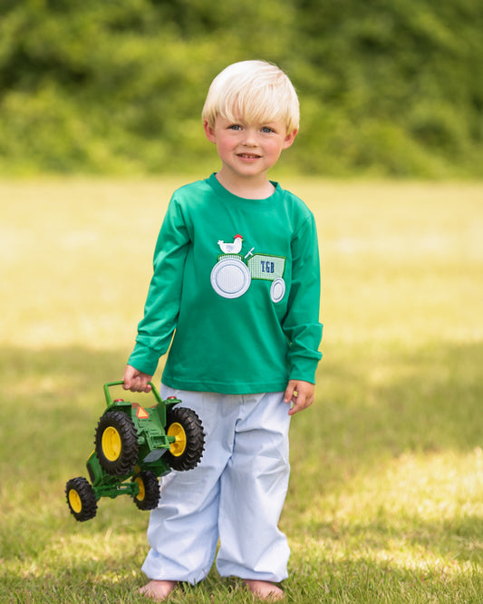 A young blonde boy stands barefoot on a grassy lawn wearing a bright green long-sleeve shirt with a white tractor graphic and light blue pants, holding a green and yellow toy tractor.