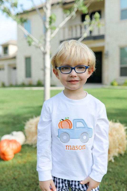 A young blonde boy wearing blue-rimmed glasses and a white long-sleeve shirt with a personalized truck graphic and the name 'mason' in orange, standing outdoors in a yard with pumpkins and a house in the background.