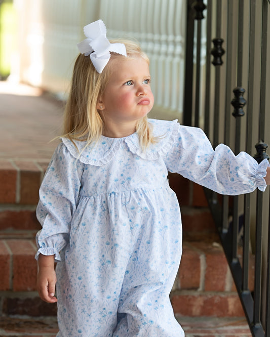 A young blonde girl wearing a light blue and white floral print baby jumpsuit with a ruffled Peter Pan collar and matching long sleeves, accessorized with a white bow in her hair. She is standing on brick stairs next to a black metal railing.
