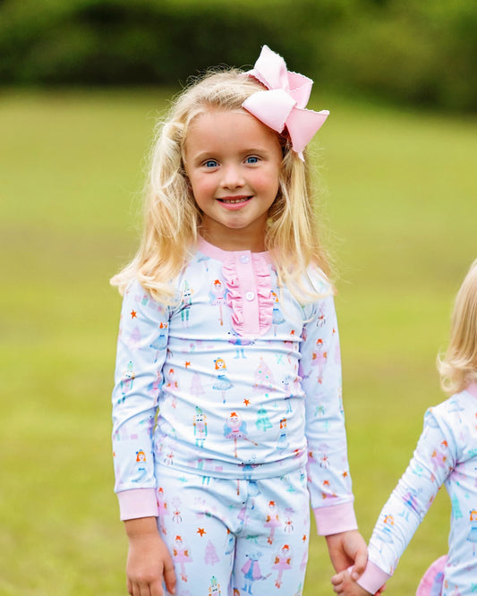 A young blonde girl wearing a light blue long-sleeve pajama set with a pink bow in her hair, standing in a grassy outdoor setting.