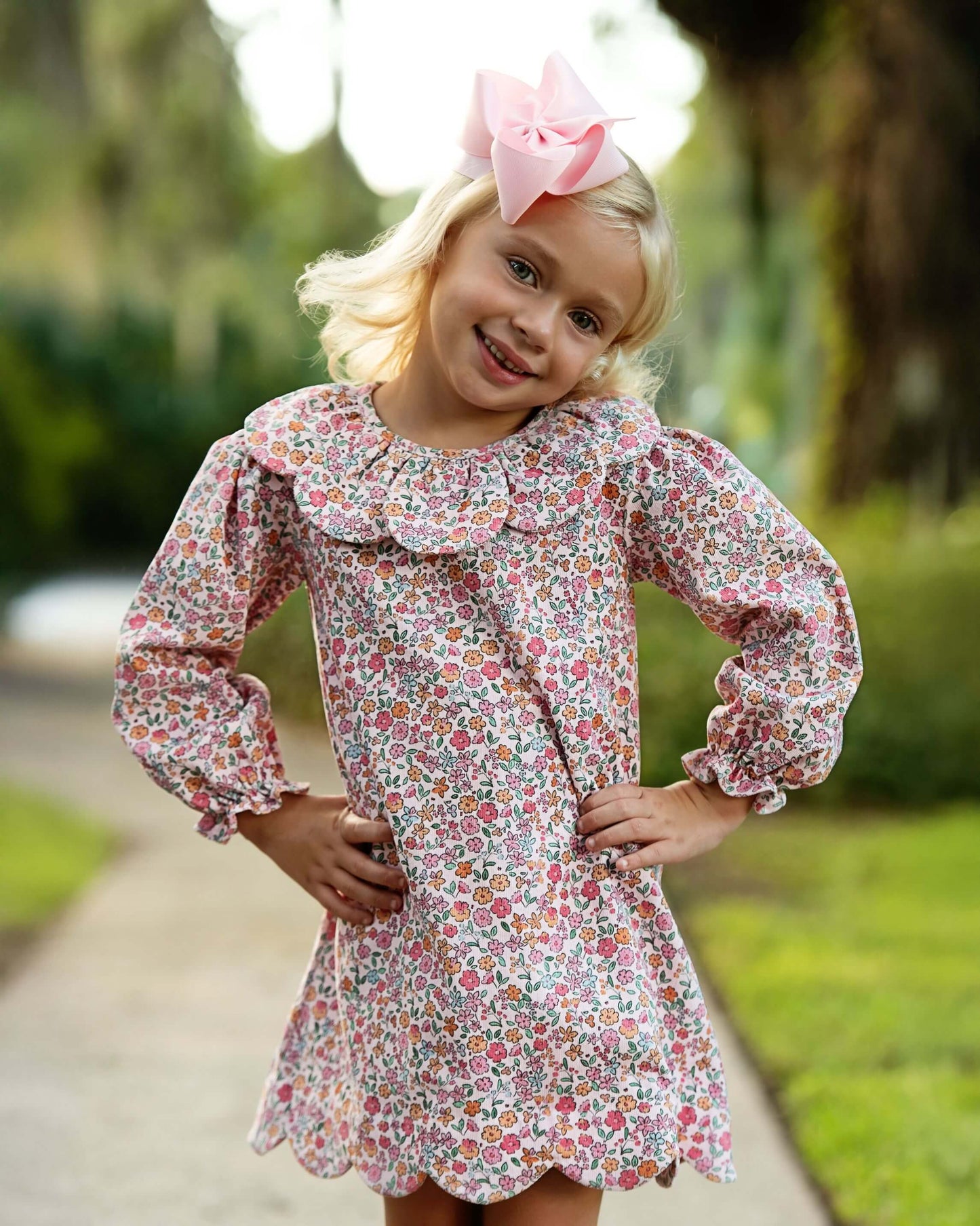 A young blonde girl wearing a pink and white floral print long-sleeve dress with a ruffled collar and a matching pink bow in her hair, standing outdoors on a path with trees in the background.