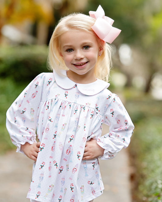 A young blonde girl wearing a white long-sleeve dress with a pink and blue floral print, a white Peter Pan collar, and a large pink bow in her hair, standing outdoors on a blurred path.