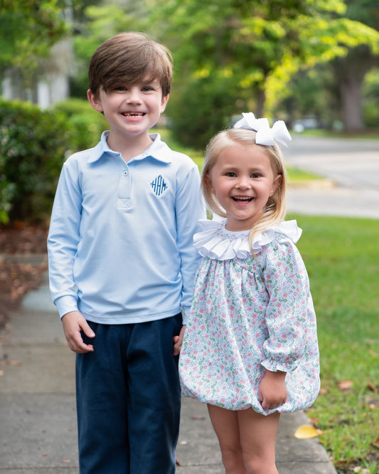 A young boy and girl standing together outdoors on a sidewalk. The boy wears a light blue long-sleeve polo shirt with dark navy pants, and the girl wears a white ruffled collar dress with a light blue and pink floral print and a white bow in her hair.