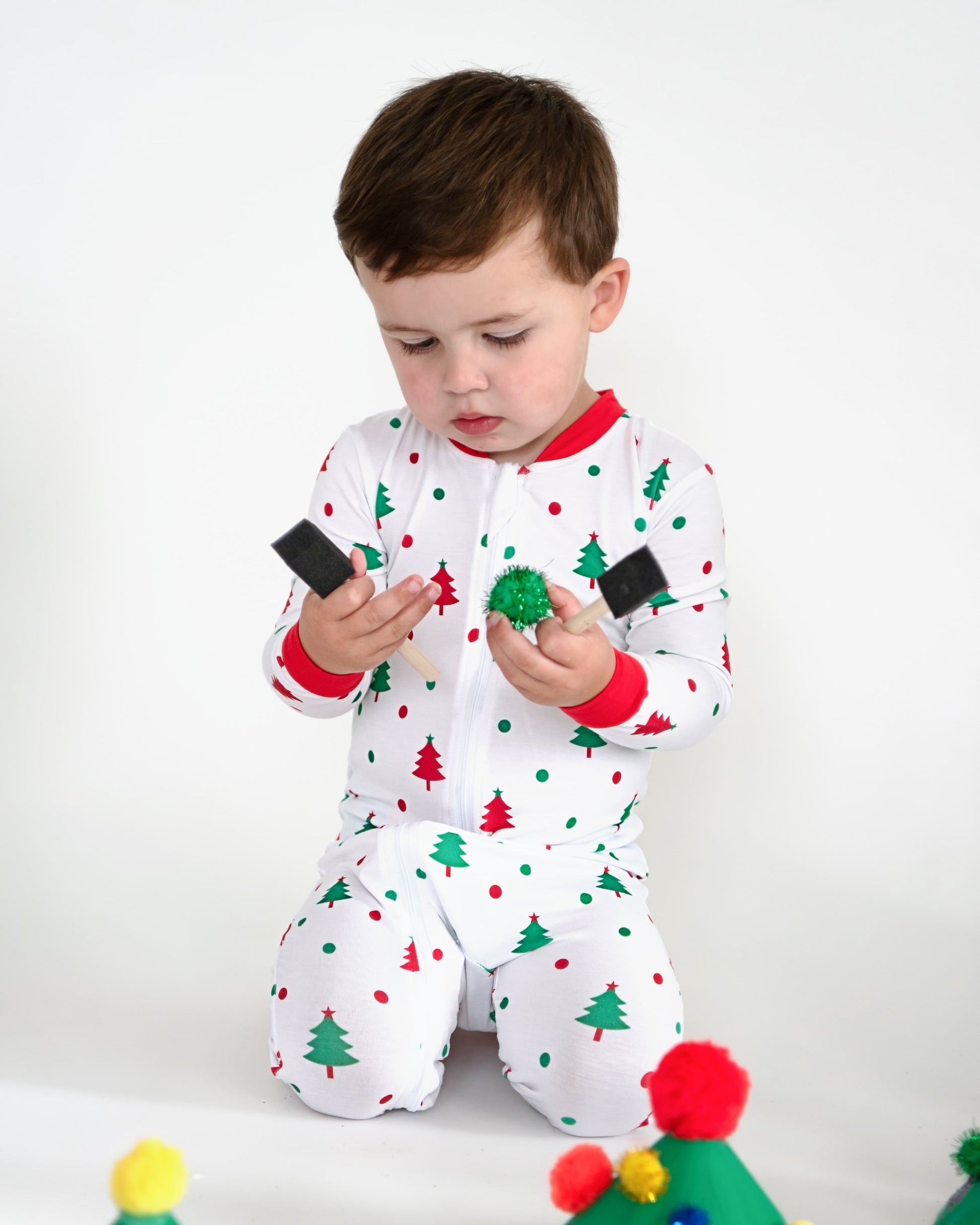 A young boy in holiday pajamas holding paint brushes.