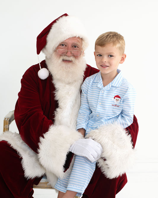 A young boy in light blue and white striped pajamas sits next to a man dressed as Santa Claus, who is wearing a red velvet suit with white fur trim. The boy has a small Santa hat patch on his pajama chest.