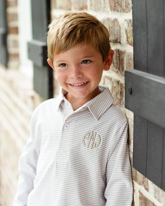 A young boy smiling and leaning against a brick wall, wearing a white long-sleeve polo shirt with thin gray stripes and a circular embroidered monogram on the chest.