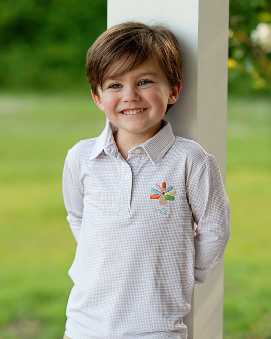 A young boy smiling and leaning against a white pillar outdoors, wearing a white long-sleeve button-up shirt with a small colorful embroidered logo on the chest.