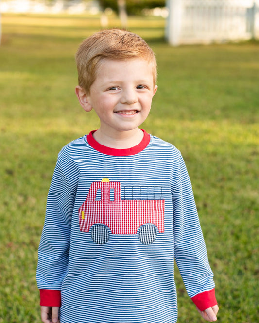 A young boy smiling outdoors on a grassy lawn, wearing a long-sleeve blue and white striped shirt with a red crew neck, red cuffs, and a red fire truck applique on the front.