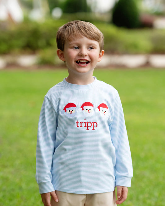 A young boy smiling outdoors on a green lawn, wearing a light blue long-sleeve shirt with three snowman faces wearing red Santa hats and the name 'tripp' in red text on the front.