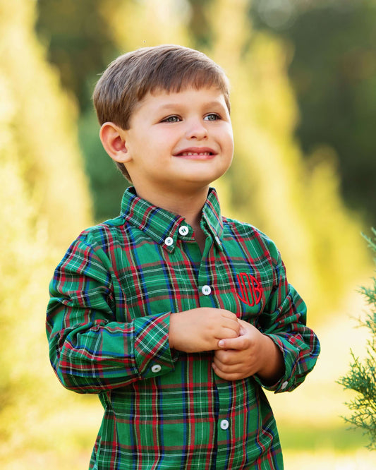 A young boy smiling outdoors, wearing a green long-sleeve plaid button-up shirt with red and white stripes and a red embroidered monogram on the chest.