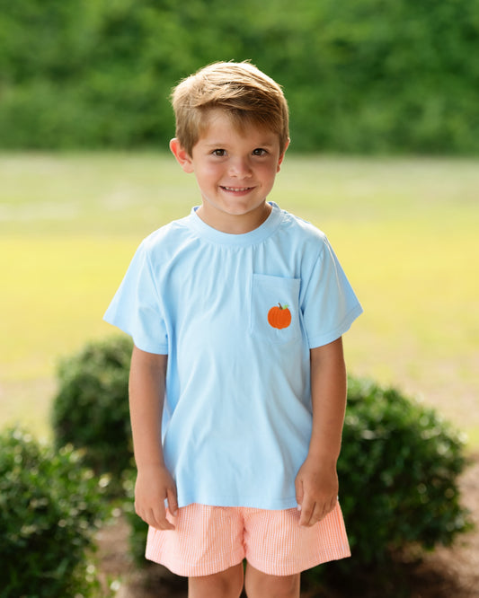 A young boy smiling outdoors, wearing a light blue short-sleeve pocket T-shirt with a small orange graphic on the chest pocket, paired with pink striped shorts.