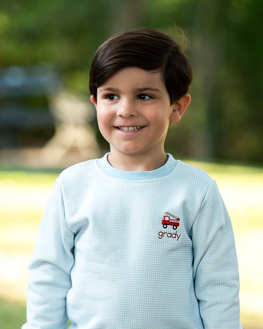 A young boy smiling outdoors, wearing a light blue waffle-knit long-sleeve shirt with a small red fire truck graphic and the name 'grady' embroidered on the chest.