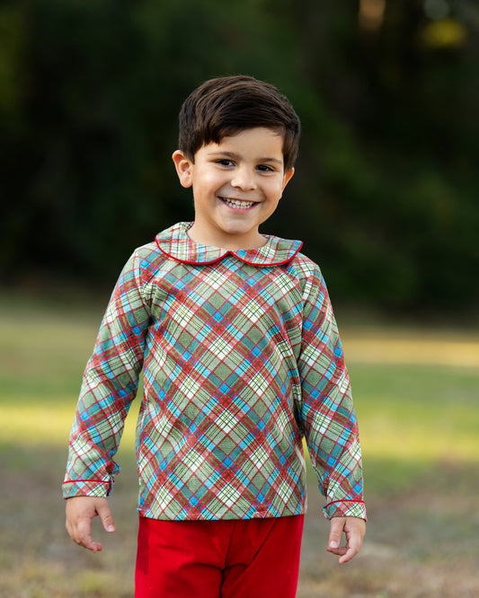 A young boy smiling outdoors, wearing a long-sleeve plaid shirt with a Peter Pan collar in beige, green, blue, and red, paired with bright red pants.