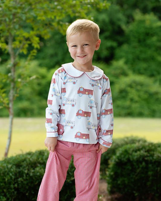 A young boy smiling outdoors, wearing a white long-sleeve pajama top with a red fire truck pattern and red-and-white gingham pants.