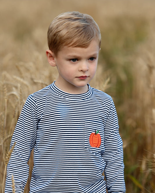 A young boy standing in a field of dry golden grass, wearing a navy and white horizontally striped long-sleeve shirt with a small orange pumpkin applique on the chest.