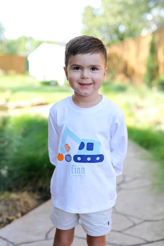 A young boy standing outdoors on a stone path wearing a white long-sleeve t-shirt with a blue excavator graphic and the name 'finn' printed on the front, paired with white shorts.