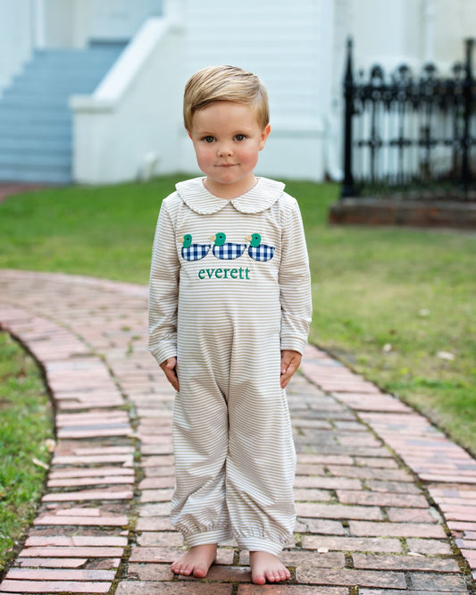 A young boy stands barefoot on a brick path outdoors, wearing a cream and white striped long-sleeve romper with a white Peter Pan collar. The front features a blue and white gingham crocodile graphic with the name 'everett' and three green alligator heads embroidered below it.
