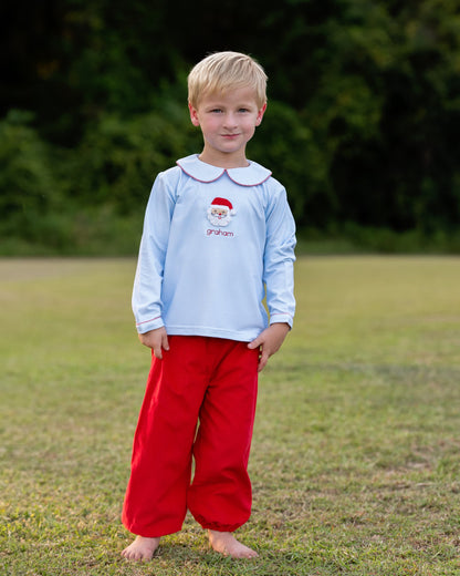 A young boy stands barefoot on a grassy field with trees in the background, wearing a light blue long-sleeve shirt with a white Peter Pan collar, a small Santa graphic and the name 'graham' on the chest, and bright red pants.