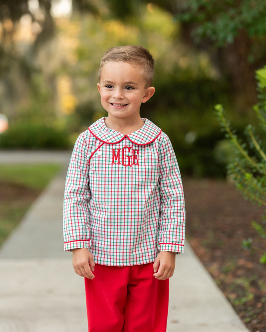 A young boy stands on a paved path outdoors wearing a long-sleeve red, green, and white gingham shirt with red trim and the letters 'MGE' embroidered in red on the chest, paired with bright red pants.