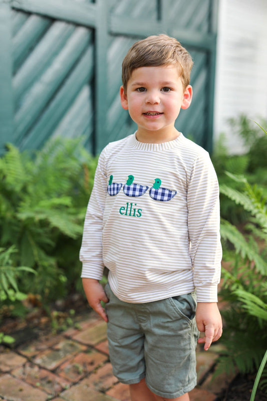 A young boy wearing a cream and white striped long-sleeve shirt with a blue and white gingham duck graphic and the name 'ellis' in green, paired with olive green shorts, standing outdoors in front of a green wooden fence and ferns.